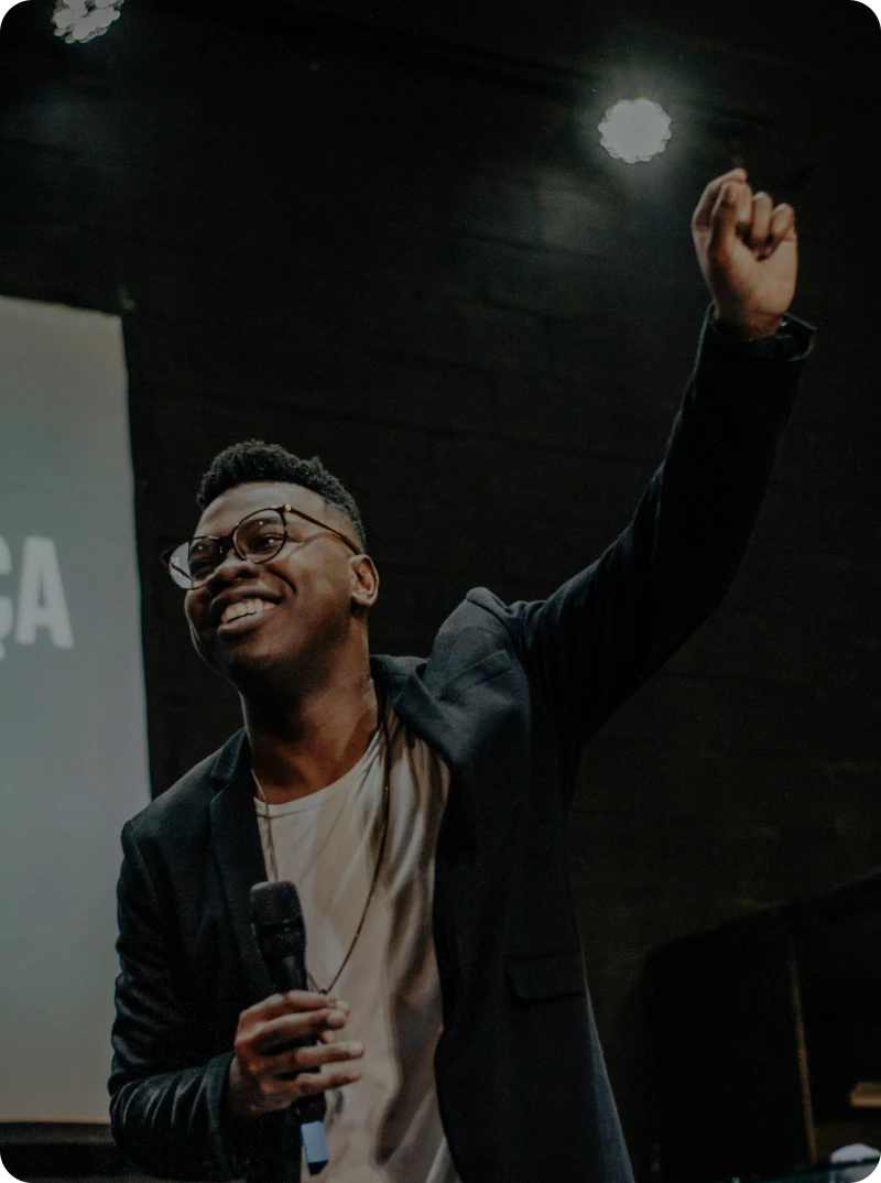 a man presenting on a stage while smiling, having a mic in one hand raising his other hand proudly